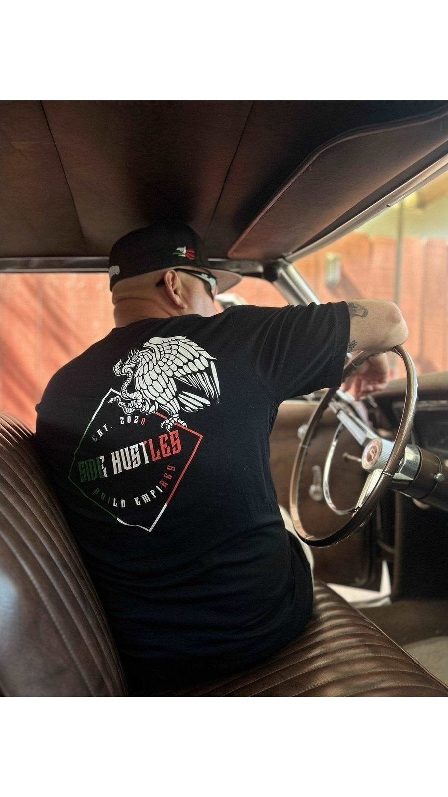 Mexican male inside of A classic car, wearing a side hustles build empires shirt. the shirt is A Bella canvas 3001 c shirt with a De-constructed Mexican flag, screen printed graphic.