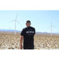 Man wearing side hustle shirt with large print on back of black shirt in front of large windmills in palm springs